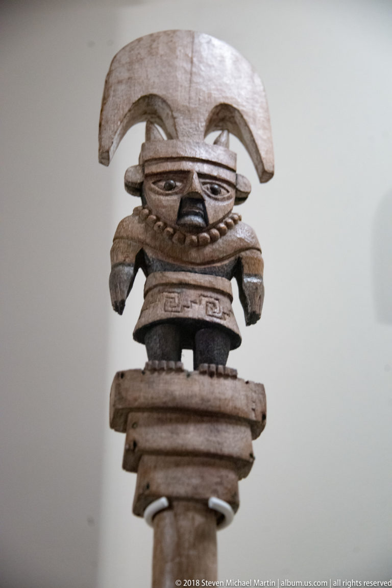 Elaborate wooden anthropomorphic figure with crescent-shaped headdress, mask-like face, projecting arms, and layered body on a post.