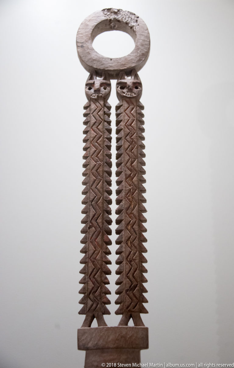 Carved wooden object with large circular top opening and two long parallel serrated strips joined near the bottom.