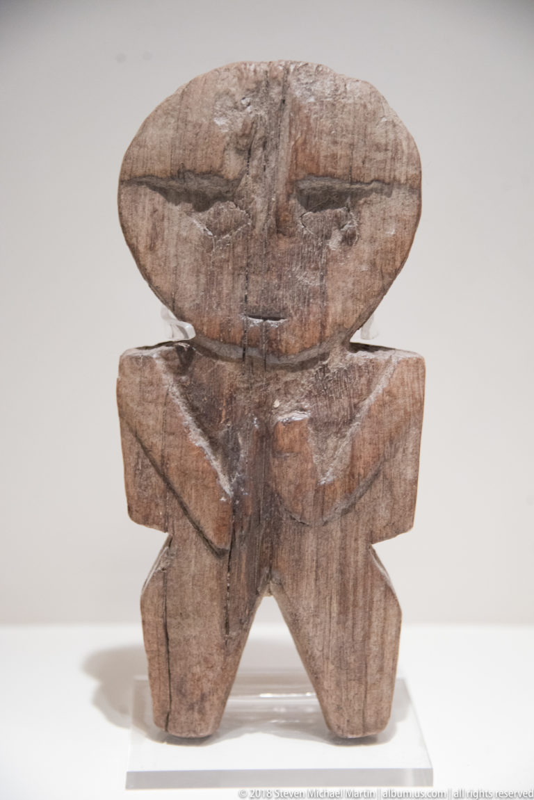 Weathered wooden anthropomorphic figure with broad head, short torso, and angular splayed legs on a museum mount.