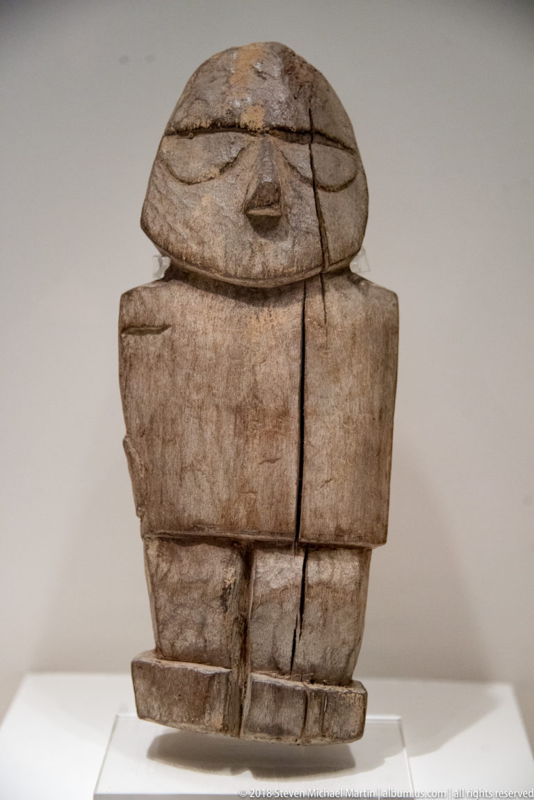 Blocky weathered wooden figure with incised eyes, small nose, rectangular torso, segmented lower body, and deep vertical cracks.
