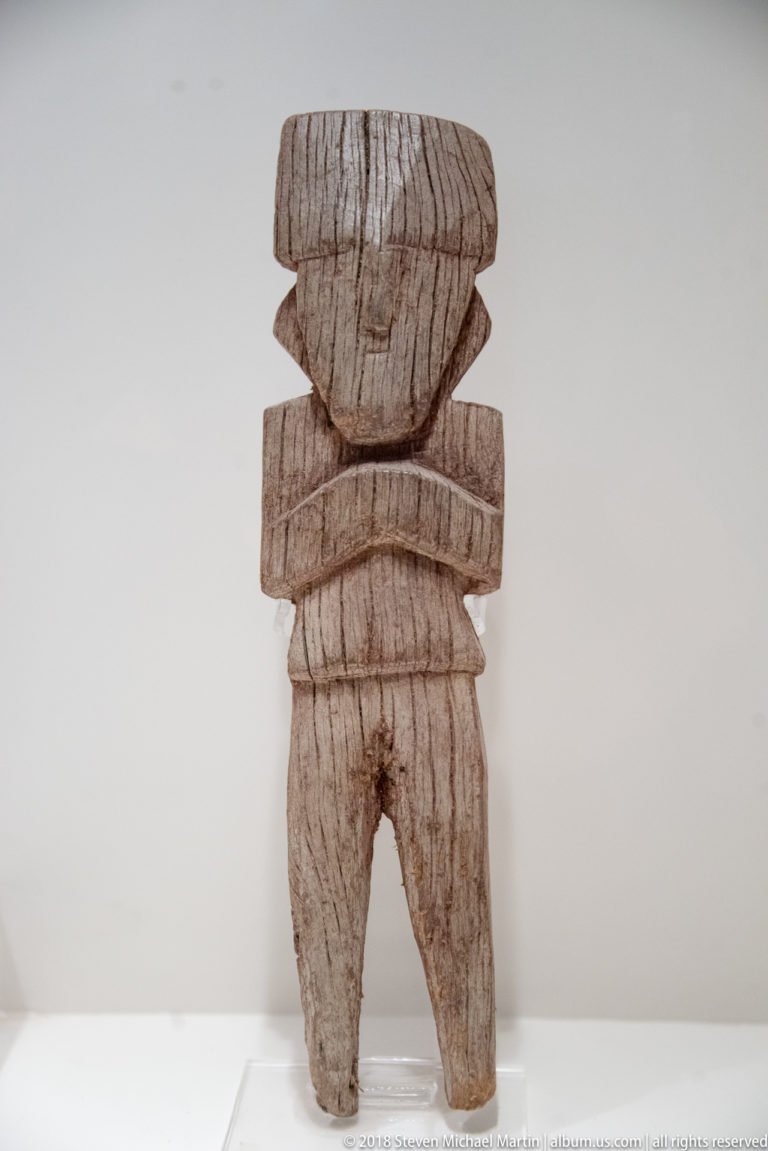 Small weathered wooden standing figure with broad head, folded arms, separated legs, and horizontal carved bands.