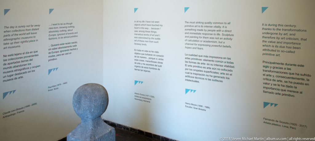 Angled museum wall with bilingual quotations and interpretive text, with a rounded sculptural head or figure in the foreground.