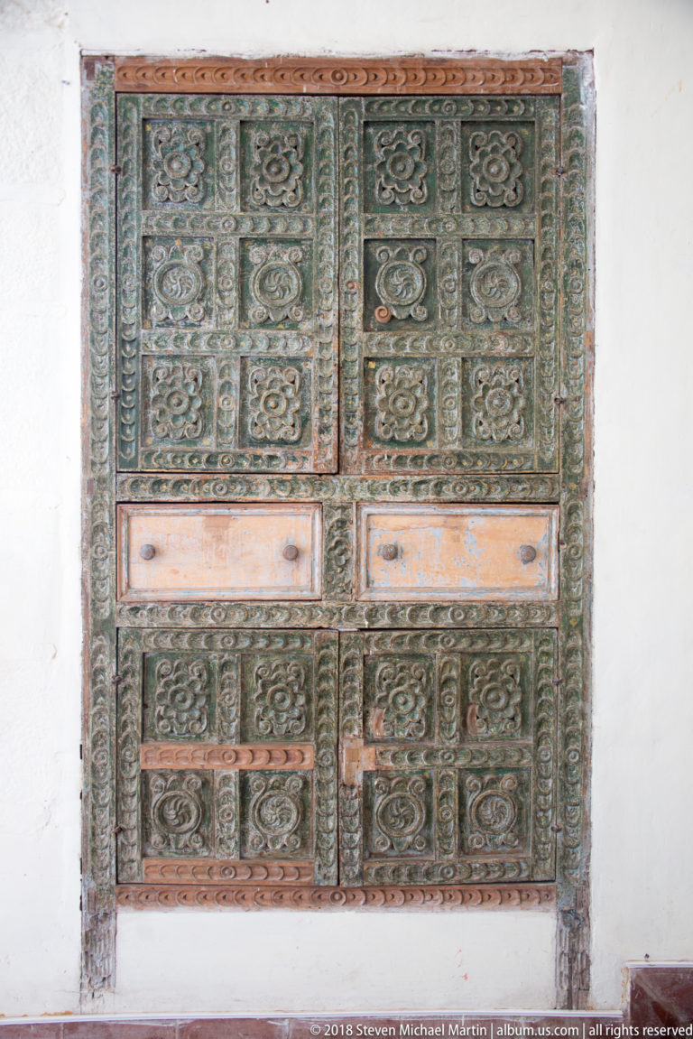 Large carved wooden door or shutter with rectangular panels filled by repeating raised floral and scroll-like motifs.