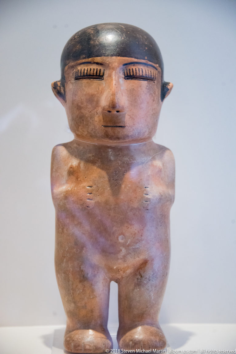 Standing pre-Columbian ceramic figure with large head, emphasized brows and ears, cylindrical torso, and short legs.