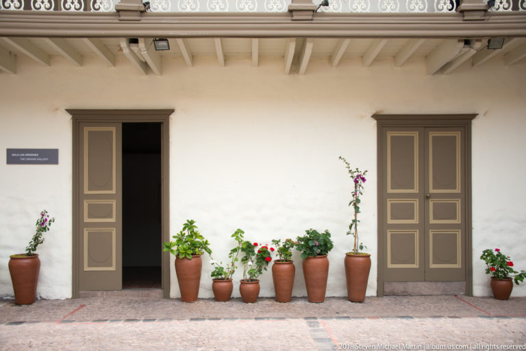 White courtyard wall with two paneled doors, an open passage, exposed ceiling beams, and a row of terracotta potted plants.