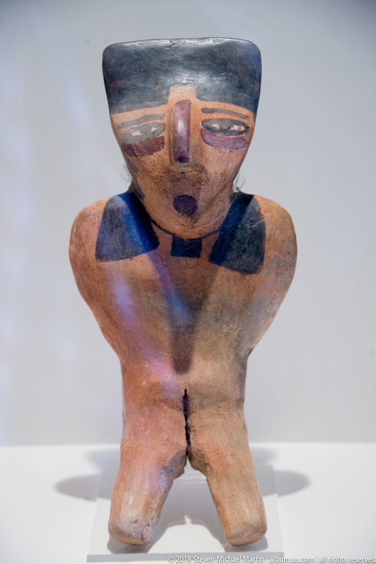 Nasca small seated ceramic figure with broad shoulders, spread legs, narrow painted eyes, projecting nose, and dark neck markings.