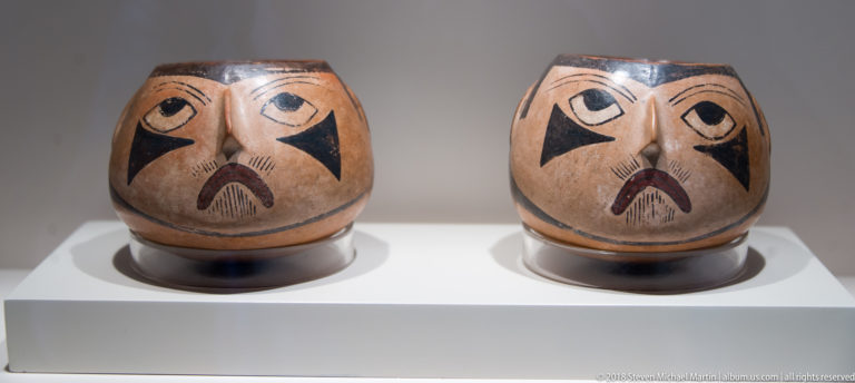 Nasca pair of tan ceramic face vessels with large eyes, downturned mouths, and black triangular markings on both cheeks.