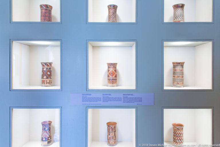 Blue-gray museum wall with illuminated square niches displaying multiple decorated pre-Columbian ceramic vessels.