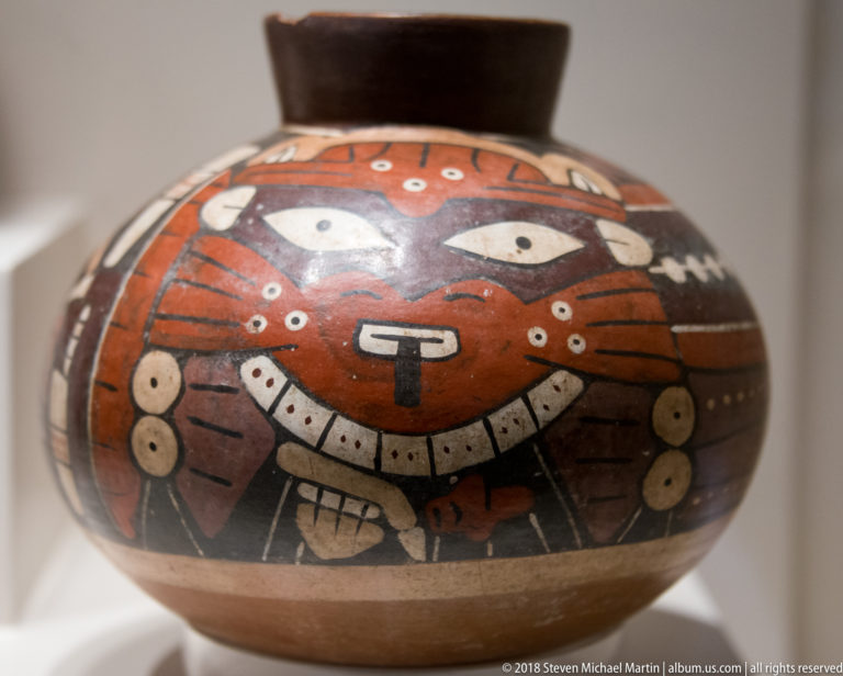 Nasca red rounded ceramic jar painted with large pale eyes, a frontal face, dotted details, and black-and-cream bands.