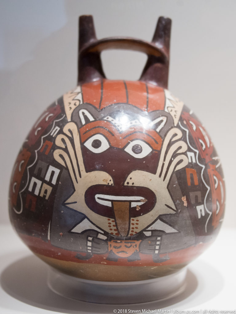 Nasca rounded bridge-handled ceramic vessel painted with a large frontal supernatural face in red, cream, black, and gray.