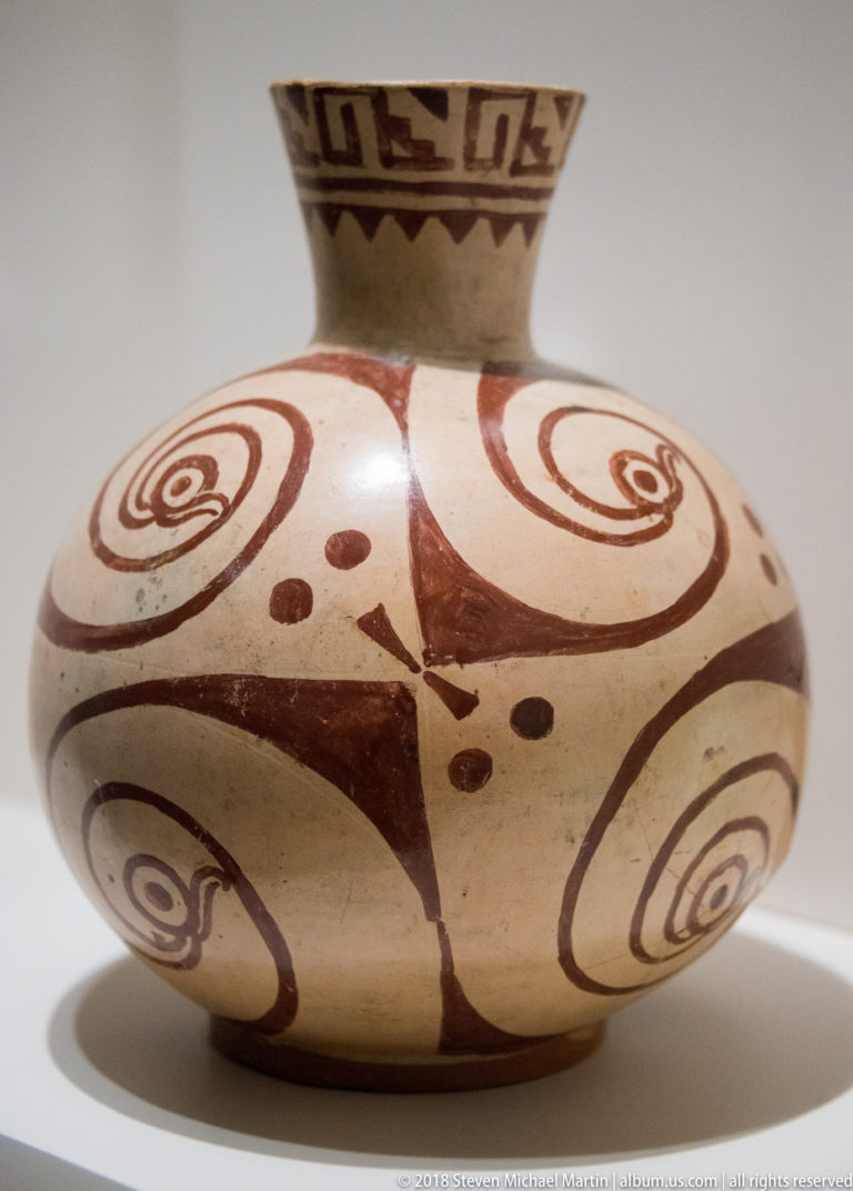 Cream globular ceramic vessel painted with large red-brown spirals, dots, and a geometric band around the neck.