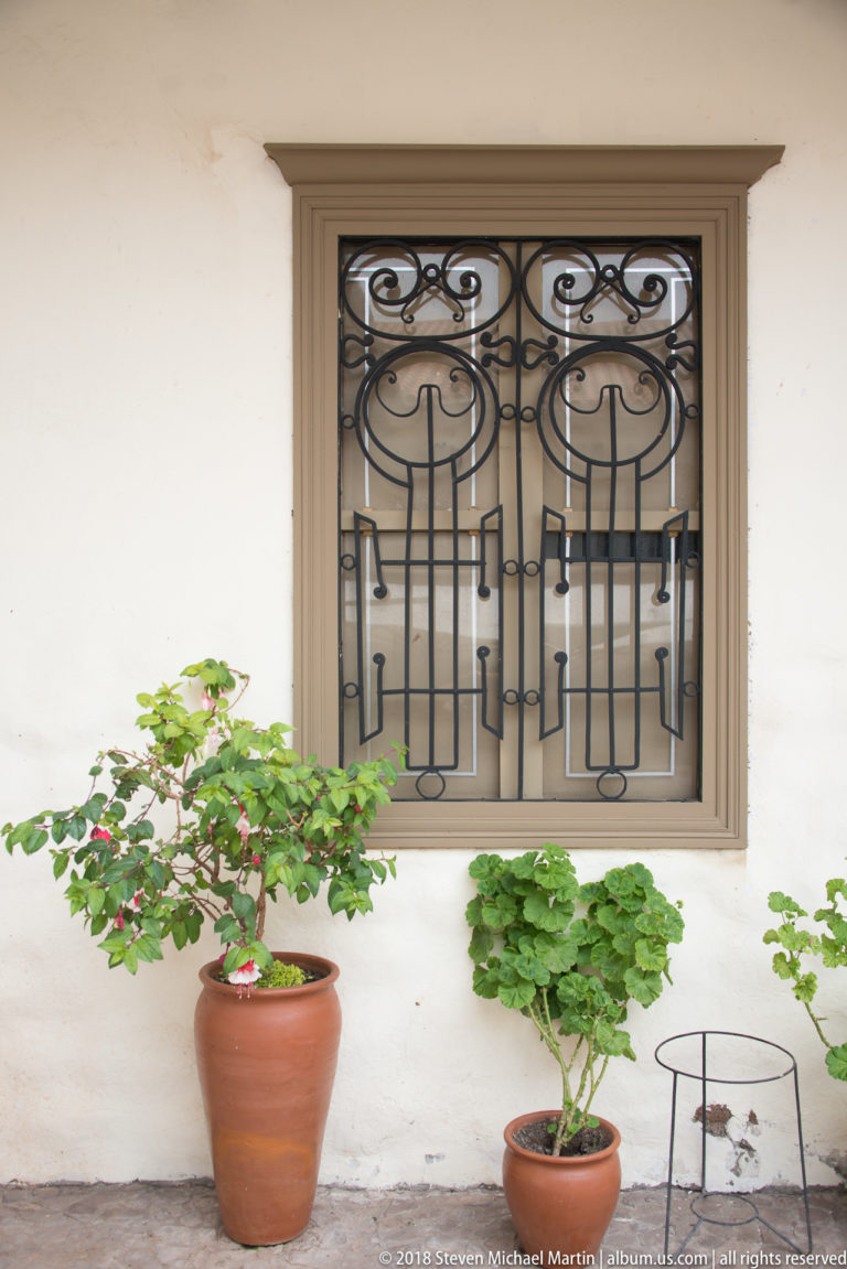 White courtyard wall with a large rectangular window, ornate black iron grille, gray surround, and terracotta pots with green plants.