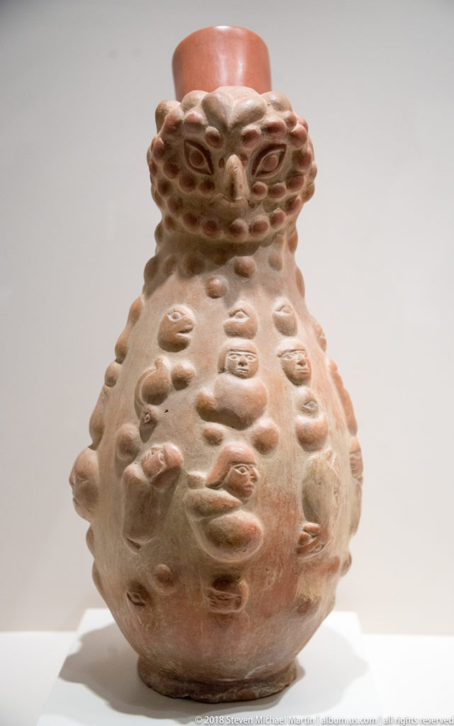 Moche globular ceramic vessel with a red cylindrical neck and numerous applied human faces and heads covering the body.