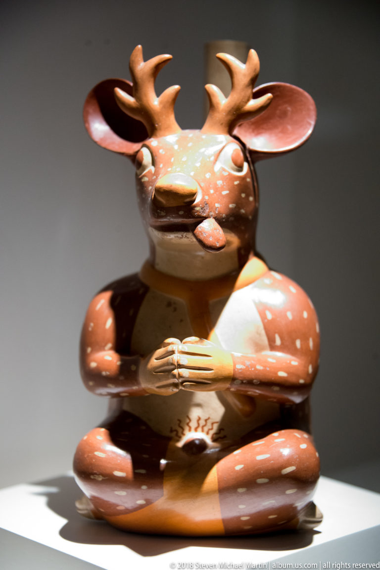 Moche ceramic effigy of an antlered spotted deer with human-like arms, hands, and a cross-legged seated posture.