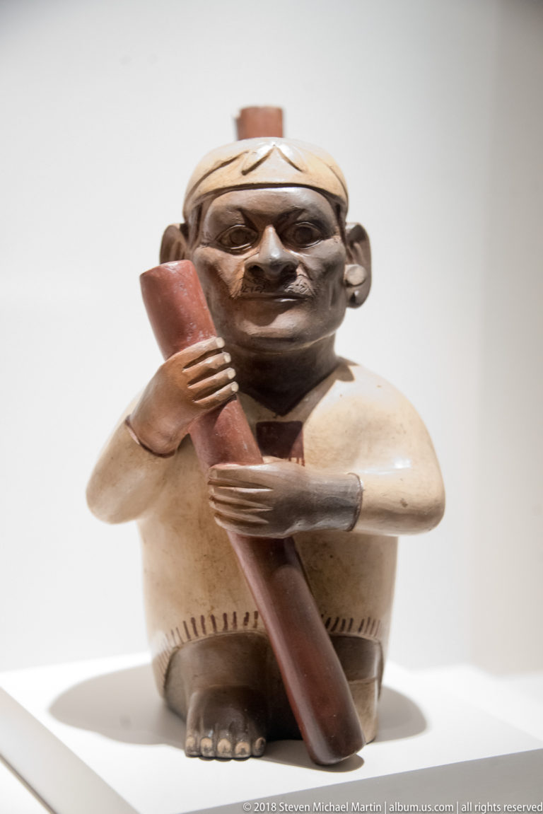 Moche seated human-effigy ceramic vessel holding a long red object across the body, with modeled face, hands, clothing, and feet.