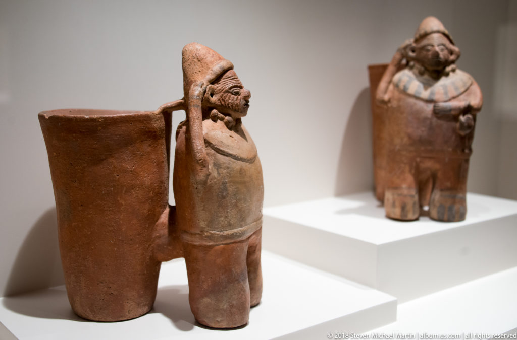 Two pre-Columbian ceramic human figures wearing conical headgear, each attached to a tall open vessel beside the body.