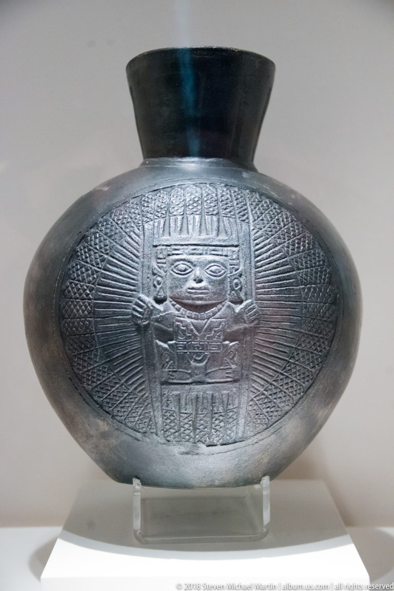 Burnished black pre-Columbian ceramic bottle with a broad body and a large incised frontal face surrounded by radiating lines.
