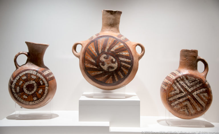 Three round pre-Columbian ceramic bottles with loop handles and bold painted geometric designs, including a central radiating face motif.