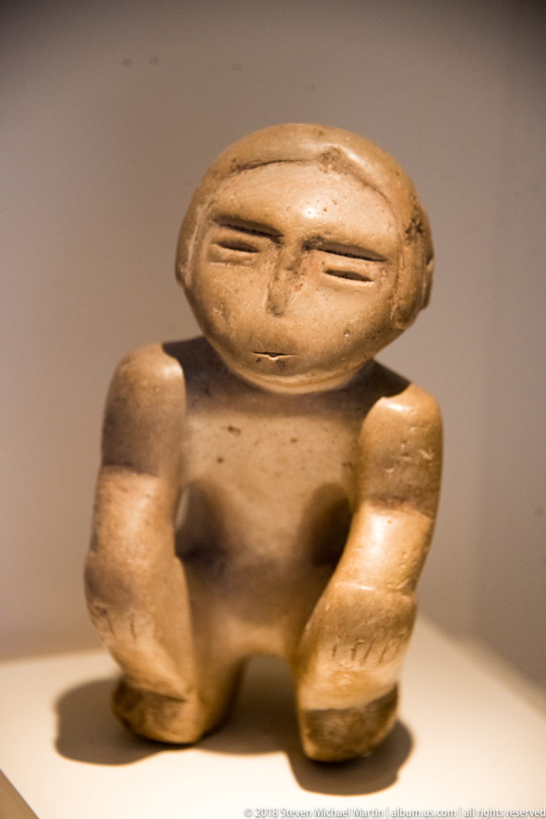 Small seated pre-Columbian ceramic human figure with broad head, narrow eyes, bent arms, and abbreviated legs.