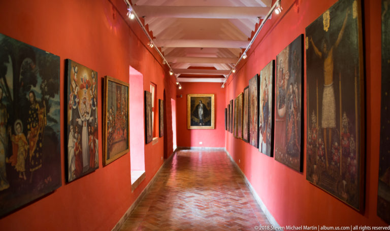 Long museum corridor with red walls, large framed paintings on both sides, track lights, and a polished patterned floor.