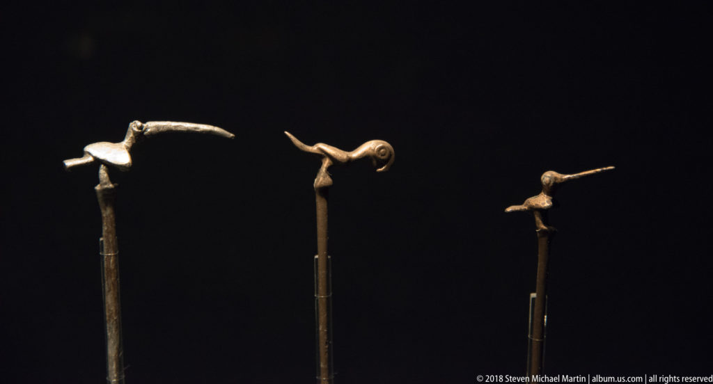Three long dark shafts topped with small metal bird figures, each shown in profile against a black background.