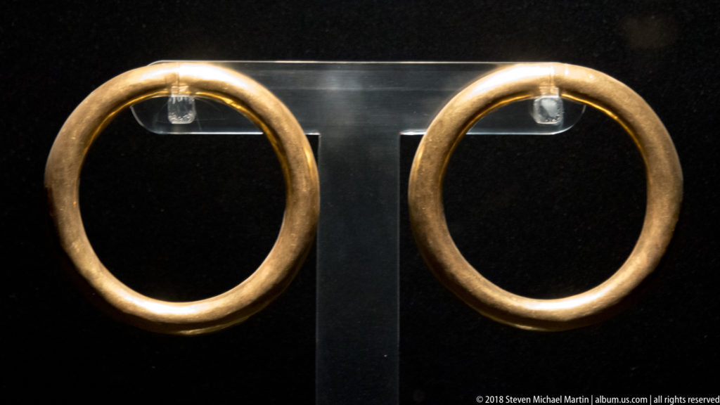 Pair of large smooth gold-colored circular hoop ear ornaments displayed side by side against black.