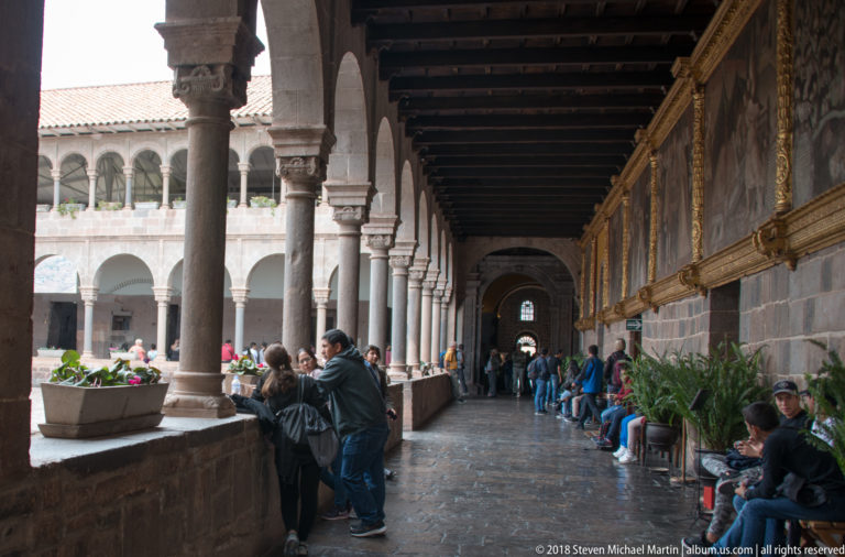 Stone-columned cloister arcade with timber ceiling, courtyard arches, and visitors seated and standing along the gallery.