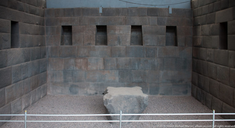Dark fitted-stone chamber with narrow wall niches and a low stone block on a gravel floor.