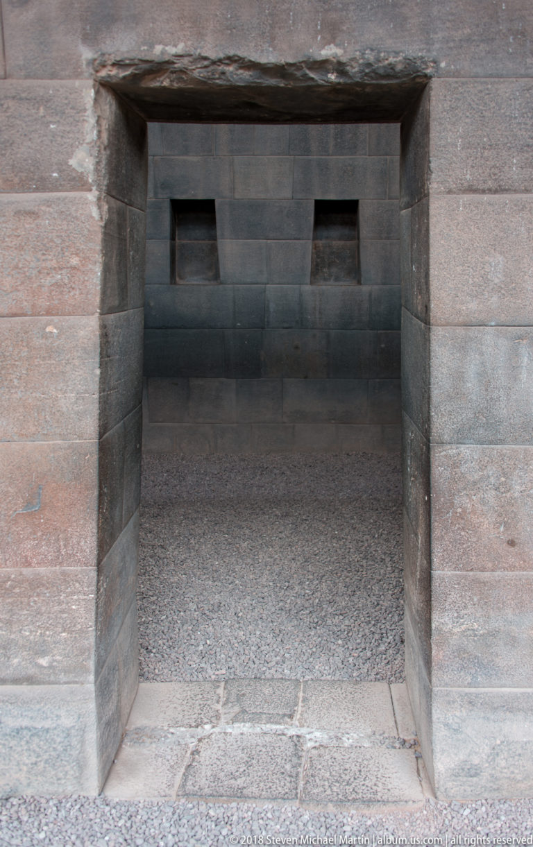 Centered view through a thick trapezoidal Inca doorway into a dark stone chamber with two small niches.