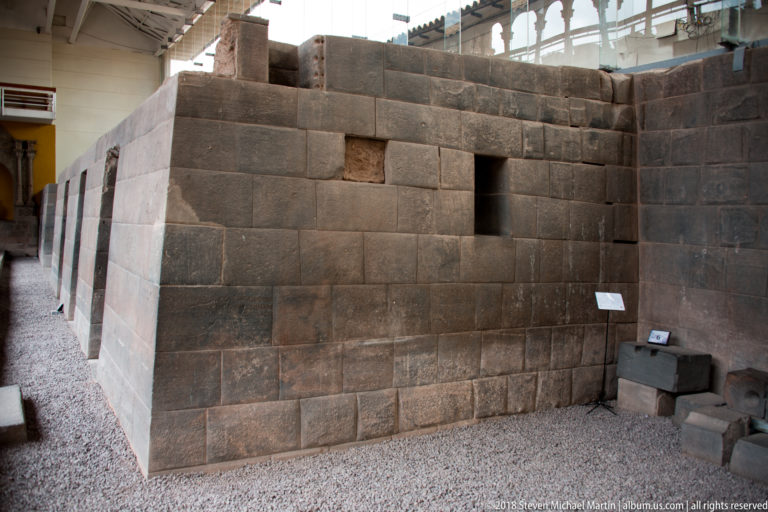 Freestanding dark Inca ashlar wall with a trapezoidal niche and doorway, surrounded by pale gravel.