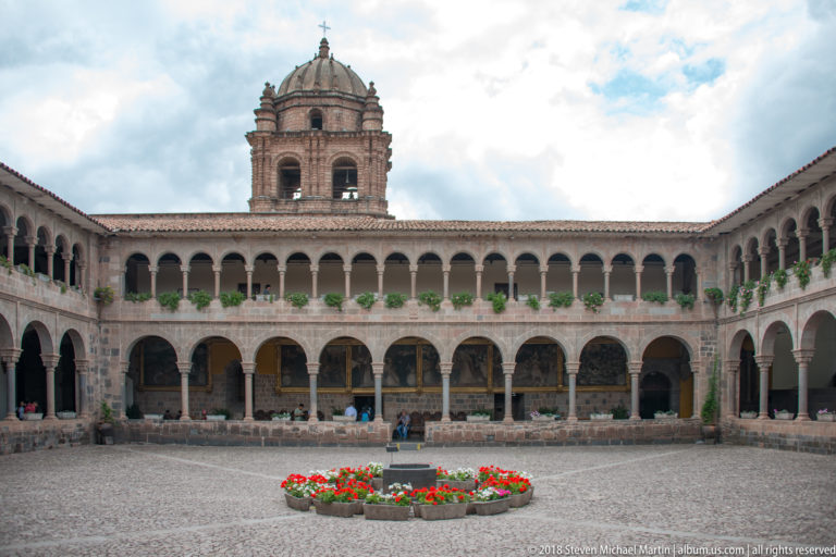 Symmetrical cobbled cloister courtyard with two-story arcades, central flower bed, low basin, and domed bell tower.