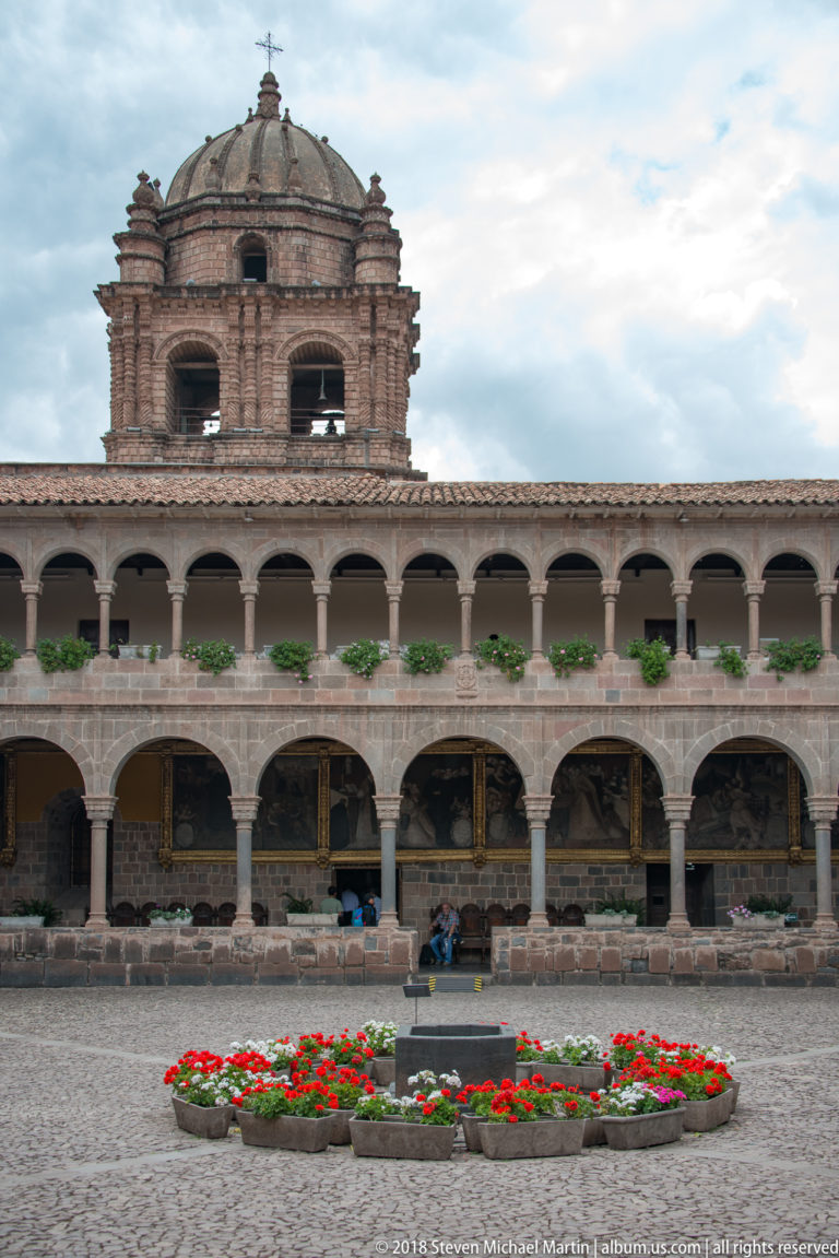 Frontal view of a two-story cloister arcade with the Santo Domingo bell tower centered above a red-and-white flower bed.