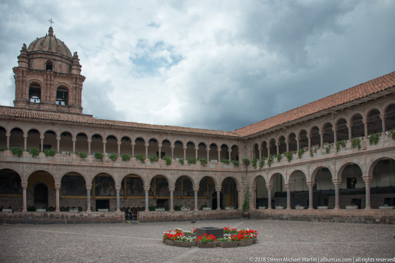 Wide Santo Domingo cloister courtyard with two-story arcades, domed bell tower, cobbles, and a circular bed of red flowers.
