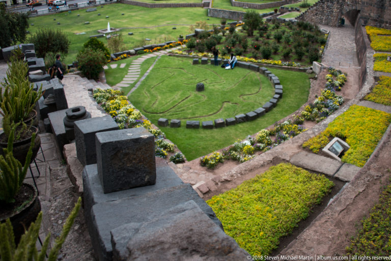 Elevated view over the circular lawn motif, flower beds, paths, and dark stone walls in the Qorikancha gardens.