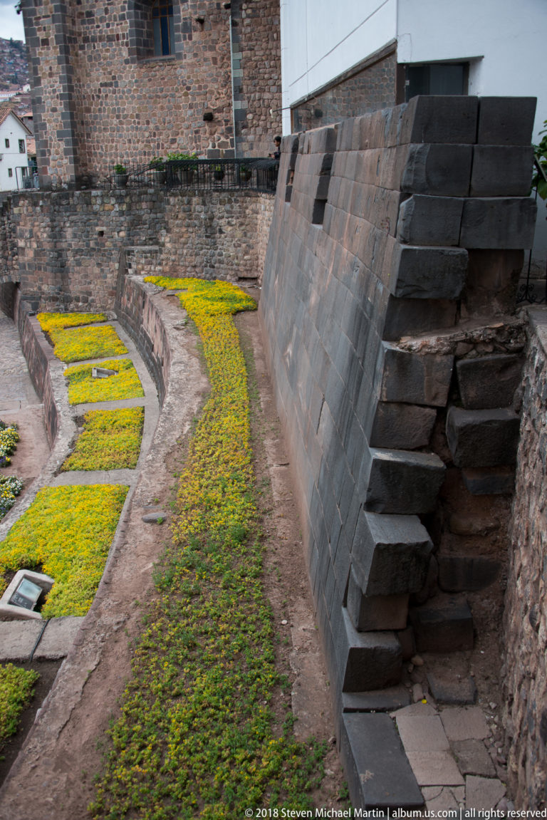 Angled end of a dark Inca stone wall above curved planted terraces with yellow-green groundcover.