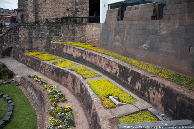Curving stepped terraces planted with yellow flowers beside a high dark Inca stone wall at Qorikancha.