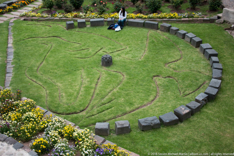 Large animal-shaped outline cut into a circular lawn, ringed by dark stones and bordered by flower beds.