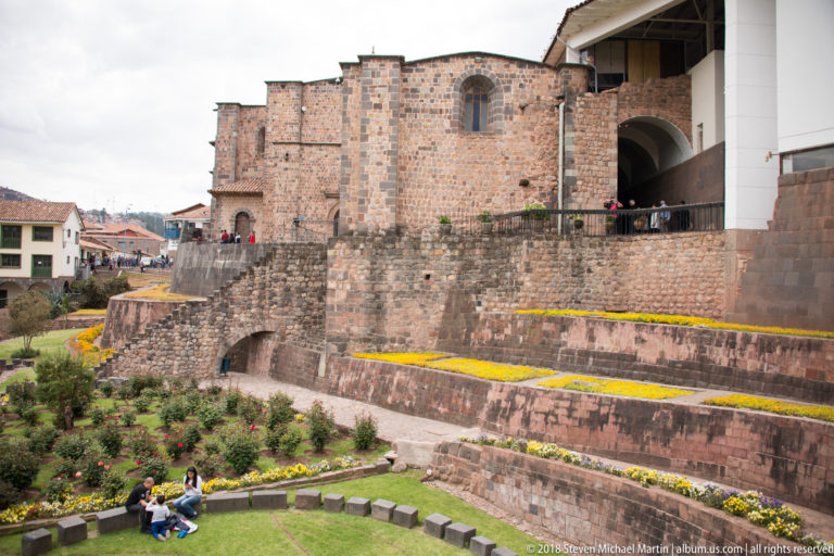 Stepped dark Inca terraces planted with flowers beneath the stone exterior of Santo Domingo, with visitors in the garden.