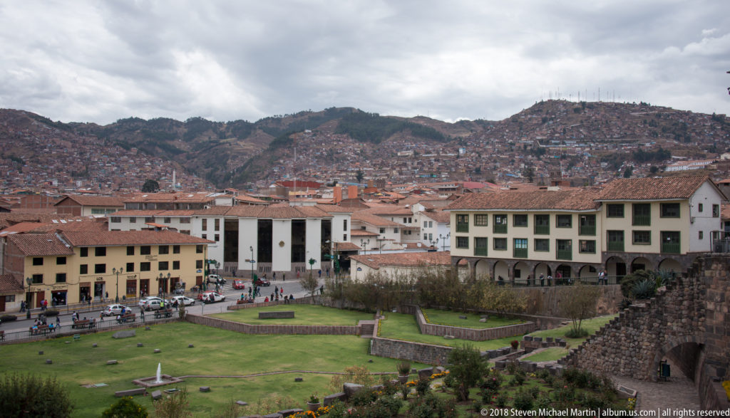 Elevated view over a green lawn and central Cusco rooftops toward densely built hills under a cloudy sky.