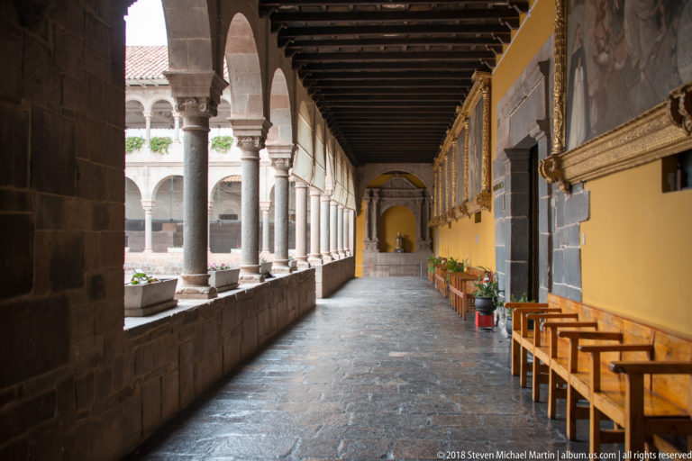 Arcaded Santo Domingo corridor with stone wall at left, religious paintings and benches along a yellow wall at right.