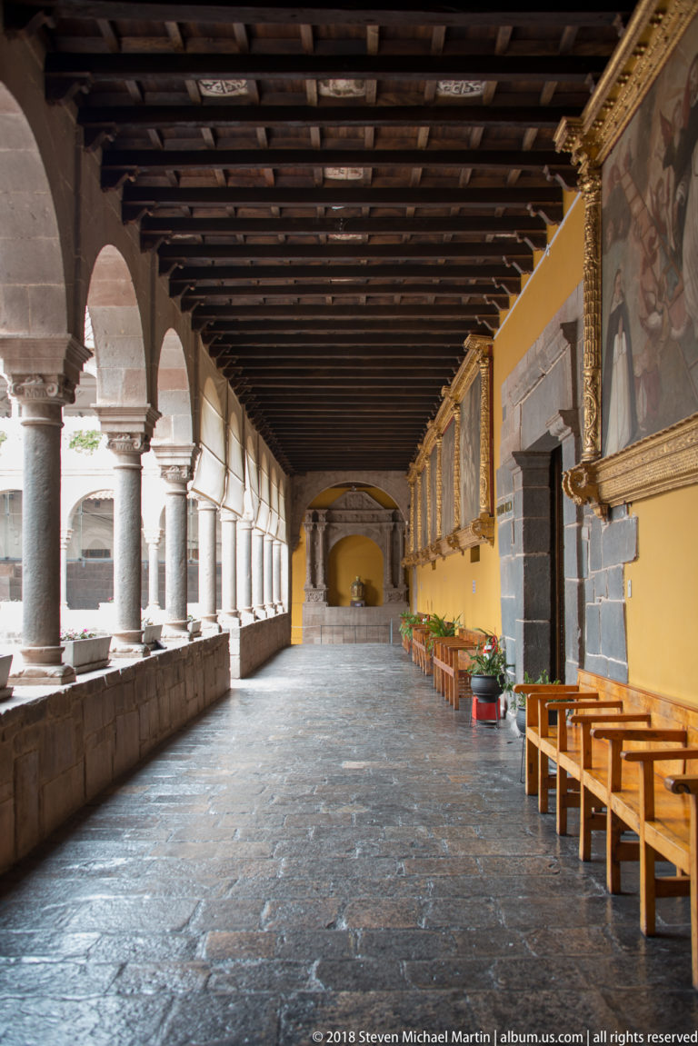 Long cloister corridor with stone arches, timber ceiling, yellow wall, framed religious paintings, and wooden benches.