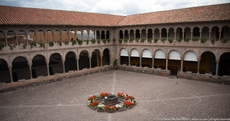 Elevated view of a cobbled cloister courtyard enclosed by two-story arcades and a circular red flower bed.