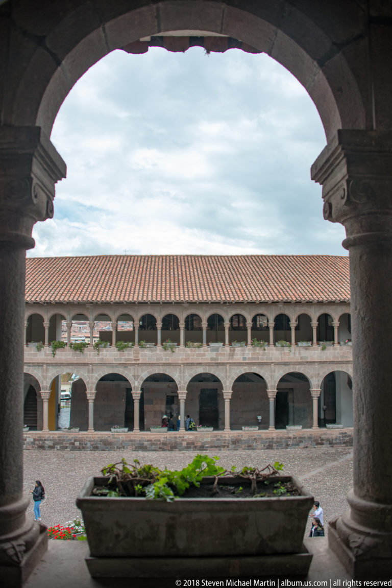 Two-story Santo Domingo cloister seen through a broad stone arch, with a rectangular planter in the courtyard.
