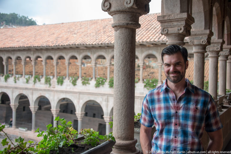 Bearded visitor in a plaid shirt beside a stone column, with the cloister courtyard and tiled roof behind.