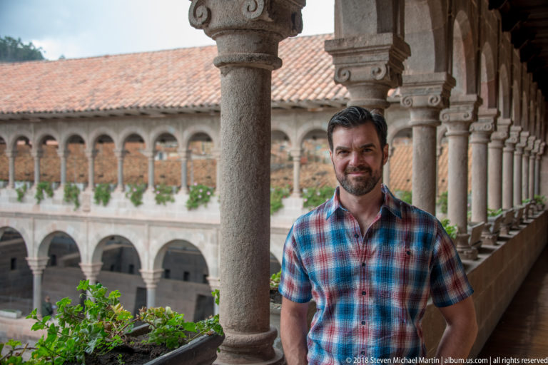 Bearded visitor in a plaid shirt standing beside carved stone columns on an upper cloister gallery.