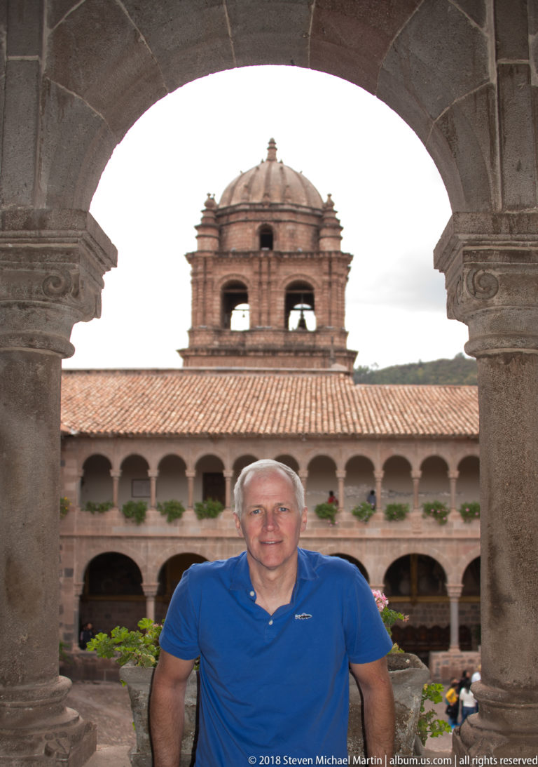 Gray-haired visitor in a blue shirt standing under a stone cloister arch with arcades and bell tower behind.