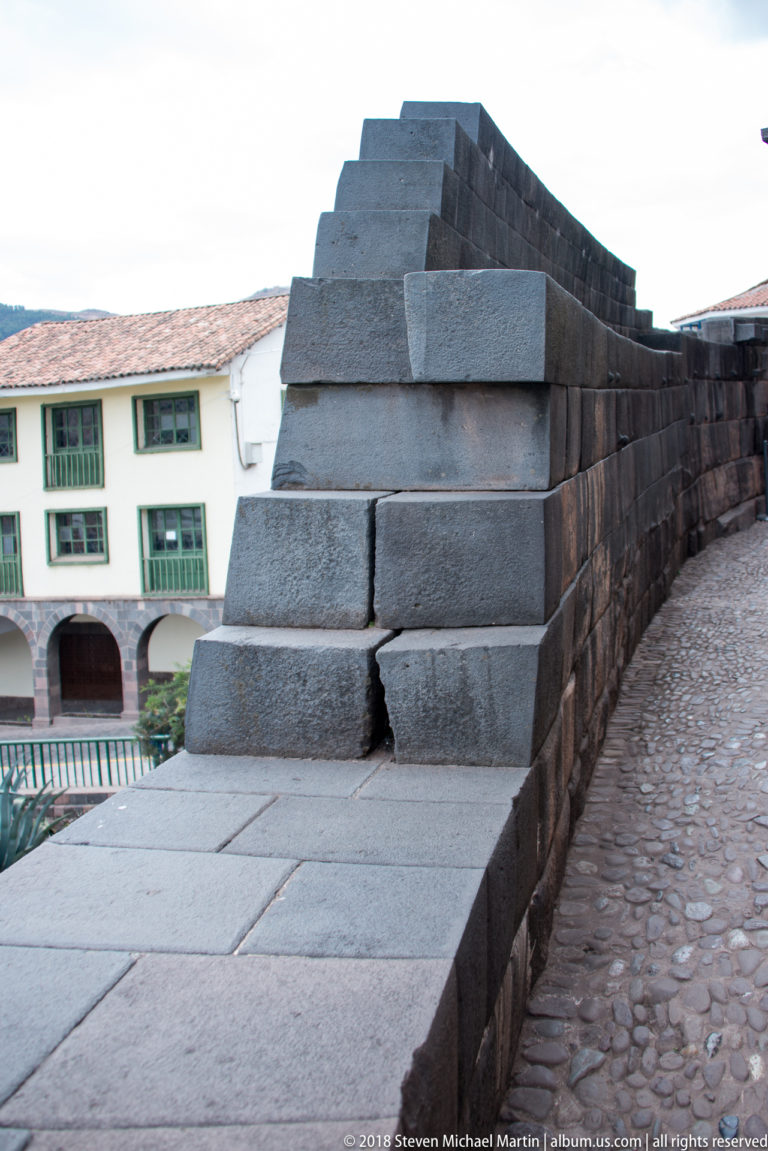 Close view along a dark Inca ashlar wall with tightly fitted stone courses beside later white buildings.