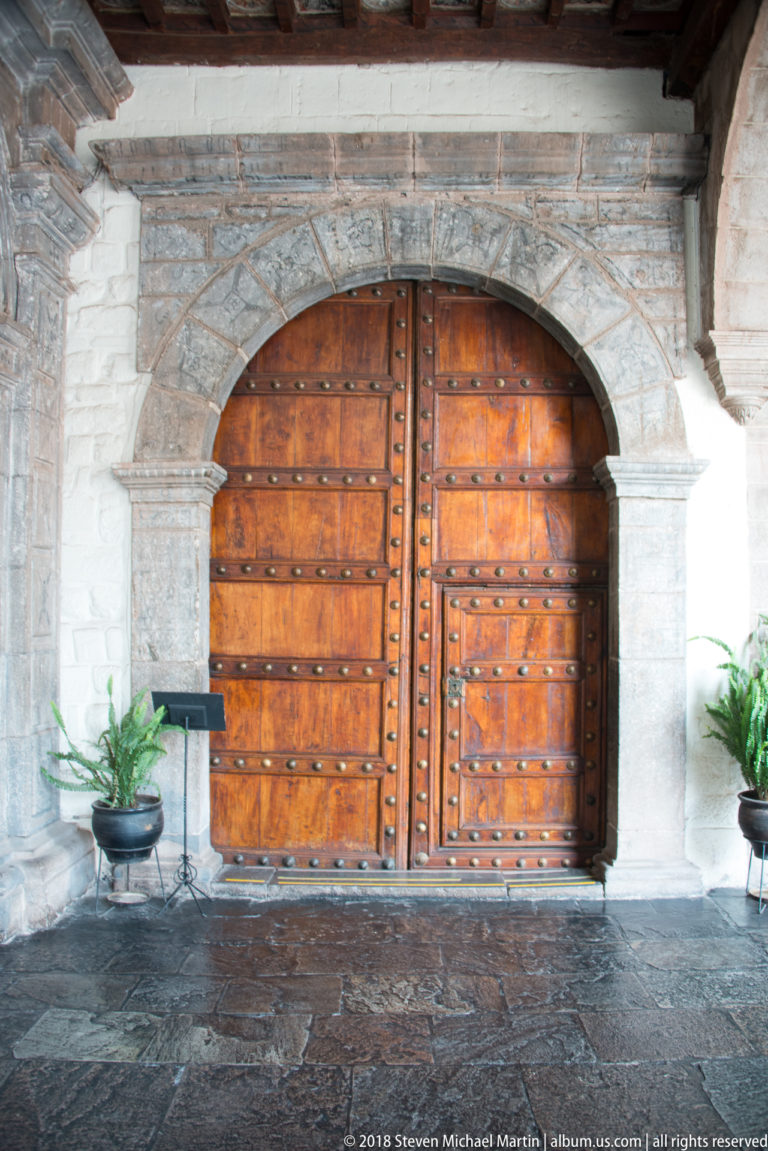 Large studded double wooden doors beneath a semicircular carved stone arch, with potted plants on either side.