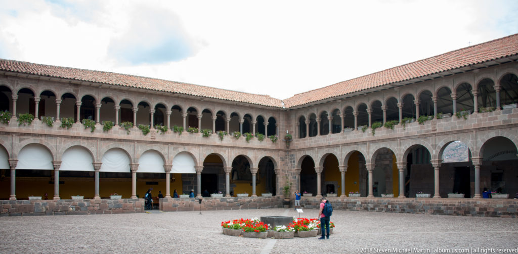 Two-story cloister courtyard with stone arcades, upper colonnade, red-tiled roofs, and visitors in the center.