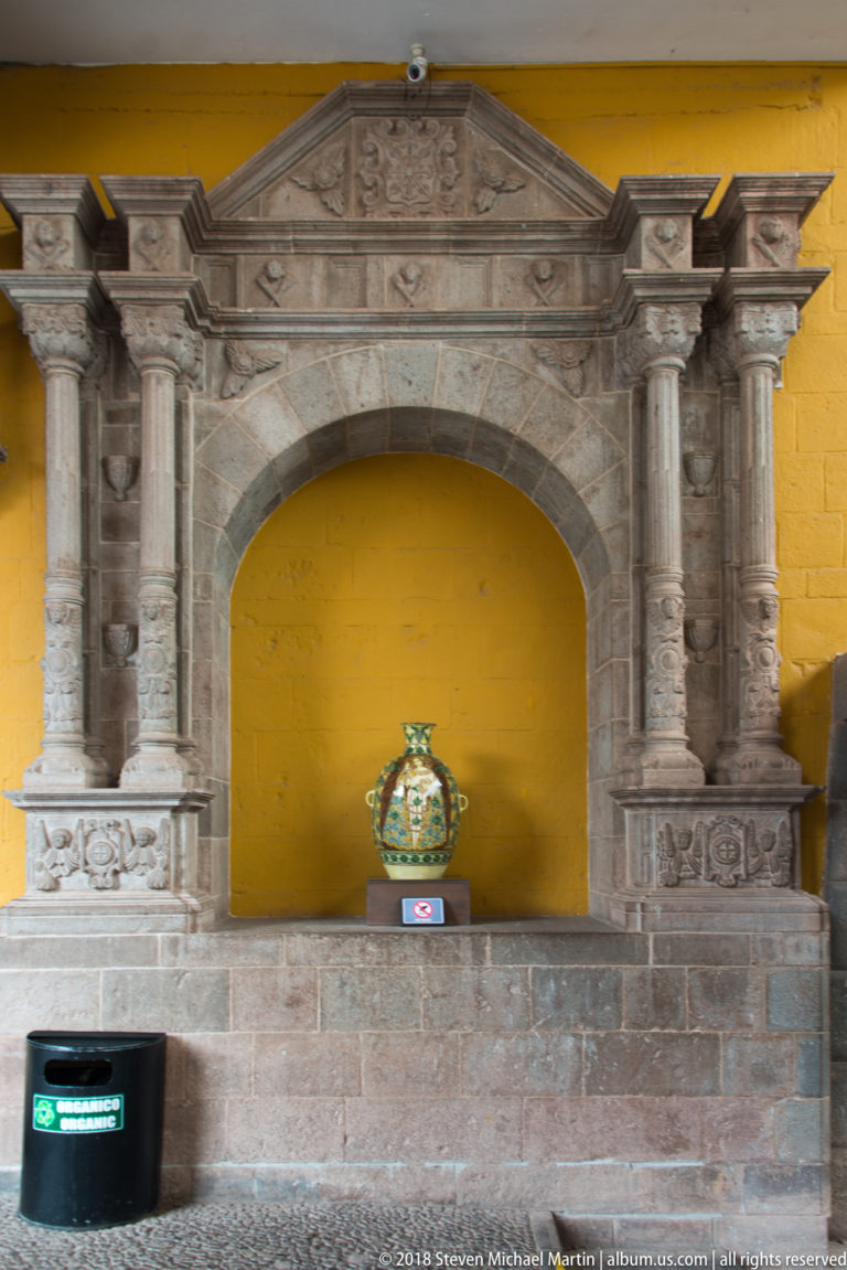 Carved stone arched niche with columns, triangular pediment, and a decorated ceramic vessel against a yellow wall.